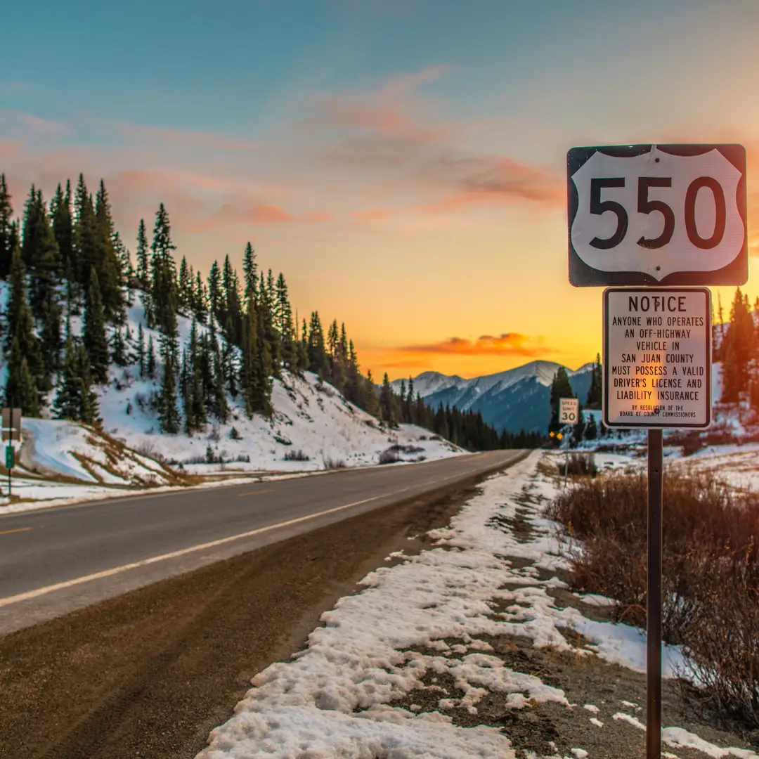 Highway 550 road sign beside a snow-covered landscape with trees, capturing the sunset sky—a scene perfect for spring adventures and outdoor fun.