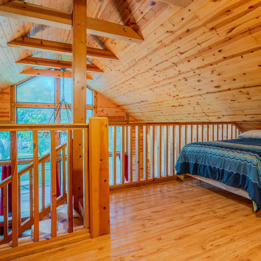 A cozy loft bedroom with wooden walls and ceiling features a blue-patterned bed and a spiral staircase, perfect for a spring break getaway. Natural light filters through the windows, offering a serene retreat similar to Lake Conroe cabins.