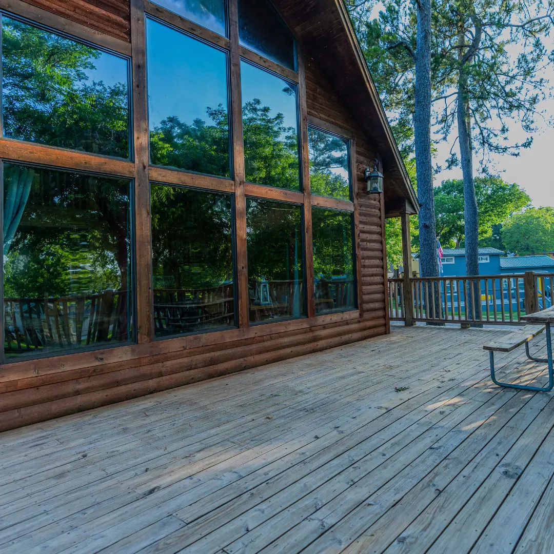 A wooden deck extends in front of a cozy Lake Conroe cabin, with large glass windows reflecting the surrounding trees. Pine trees and a partial view of another building are in the background, making it an ideal spring break getaway.