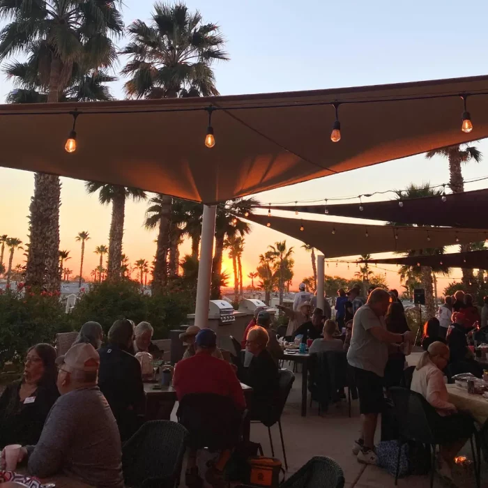 In January, people are enjoying a social gathering on a patio at sunset in Gold Canyon, surrounded by palm trees and shaded by elegant fabric canopies.