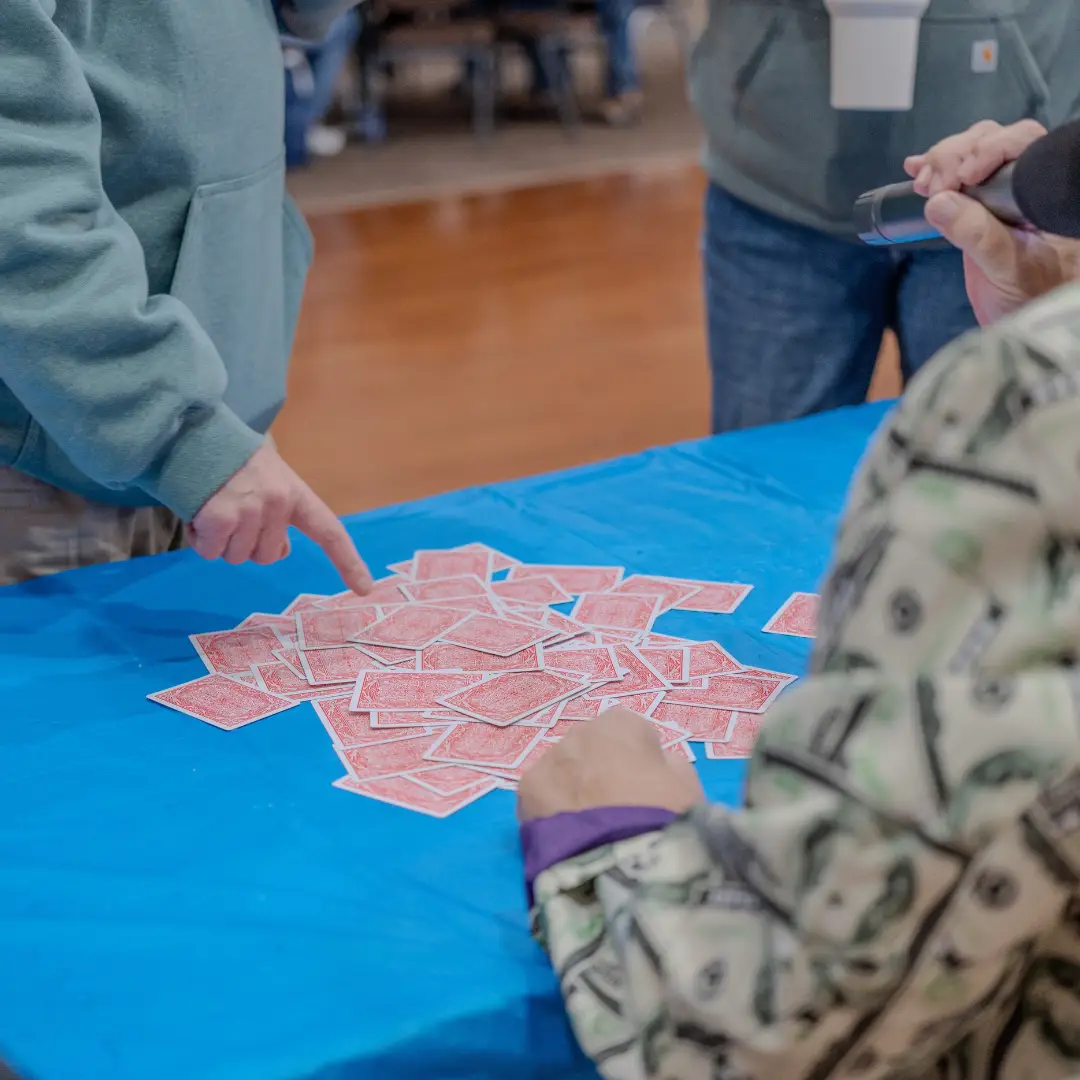 In the heart of Pueblo El Mirage, people gather around a table with scattered playing cards on a blue tablecloth. One person points at the cards, enjoying one of their favorite January activities in this vibrant community.