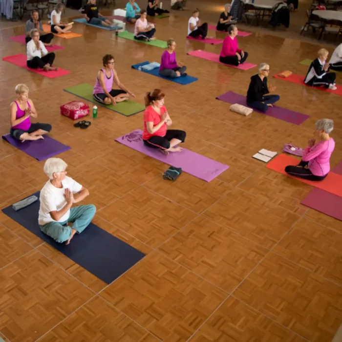 A group of winter visitors is practicing yoga on mats in a spacious indoor setting, each seated cross-legged, with wooden flooring visible.