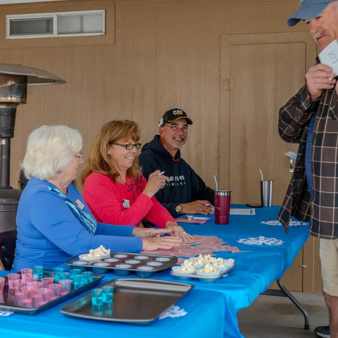 In January, three people sit at a blue table with cards and snacks in Pueblo El Mirage, assisting a standing individual. The scene is imbued with the warmth of casual, friendly activities.