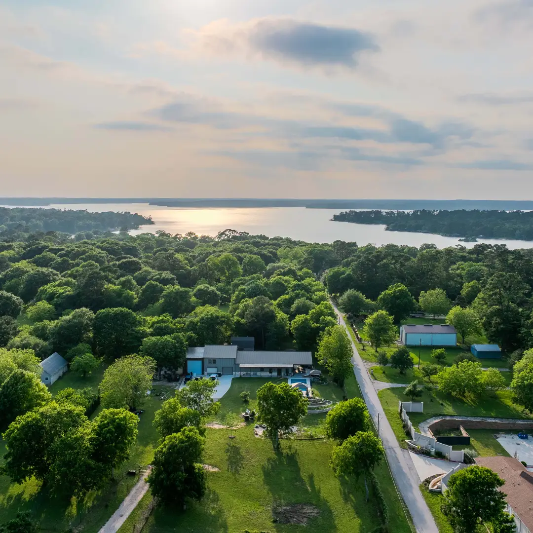 Aerial view of a rural landscape with scattered houses, a road, dense green trees, and Lake Conroe in the background under a partly cloudy sky. Perfect for an RV site or indulging in water-based activities.