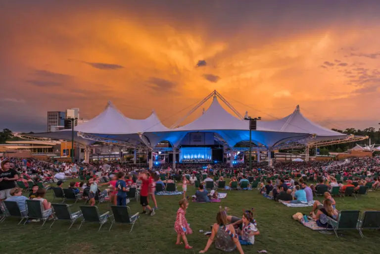 A large crowd gathers at Rayford Crossing RV Resort, an attraction known for its outdoor concert venue with a dramatic orange sky in the background. People are seated on grass and chairs, and a stage is visible under a tent-like structure, creating the best atmosphere for music lovers.