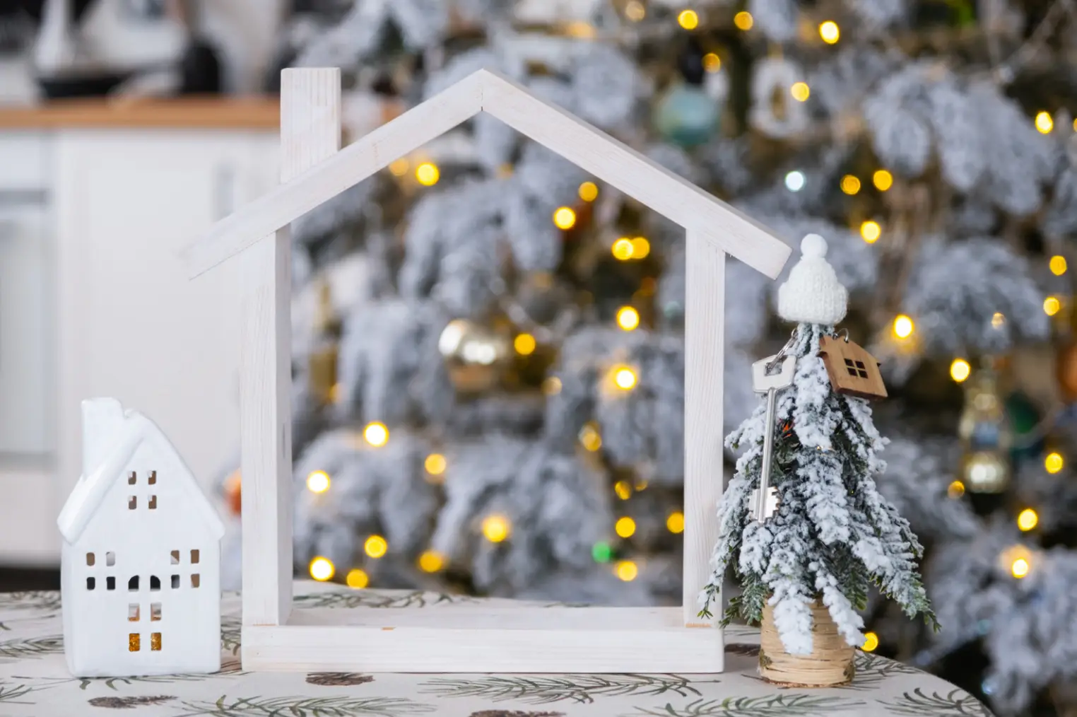 A wooden house frame and a small decorative snowy tree sit on a table, with the blurred glow of a Christmas tree bringing warmth to the holiday season. It's like finding the perfect festive hideaway amidst an RV resort in Oak Forest.