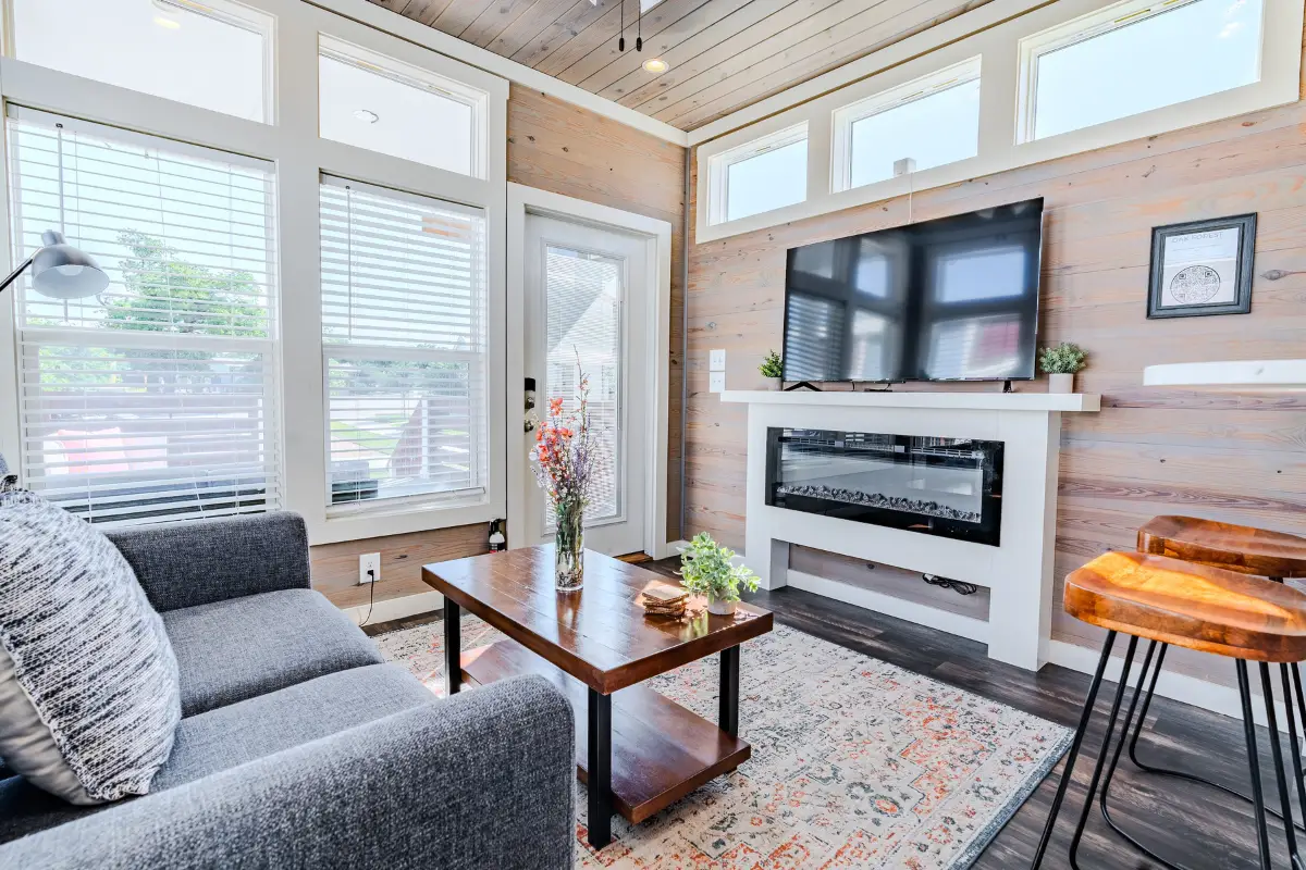 Cozy living room with a gray sofa, wooden coffee table, wall-mounted TV, and electric fireplace. Sunlight streams through large windows at Oak Forest RV Resort, highlighting a colorful rug. A perfect retreat for your holidays.