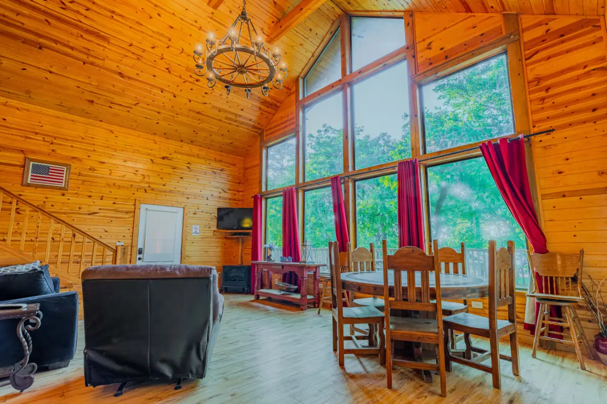 A cozy wooden cabin interior near Lake Conroe features large windows, red curtains, a dining table, leather chairs, and a chandelier. An American flag proudly decorates the wall, making it perfect for memorable holidays.