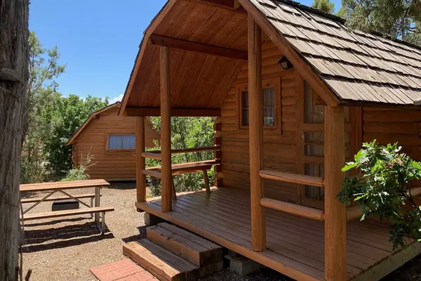Two charming wooden cottages with shingled roofs nestle in a wooded area at Oasis RV Resort, Durango, Colorado. One boasts a small porch where you can relax while the nearby picnic table invites you to enjoy meals under a clear blue sky.