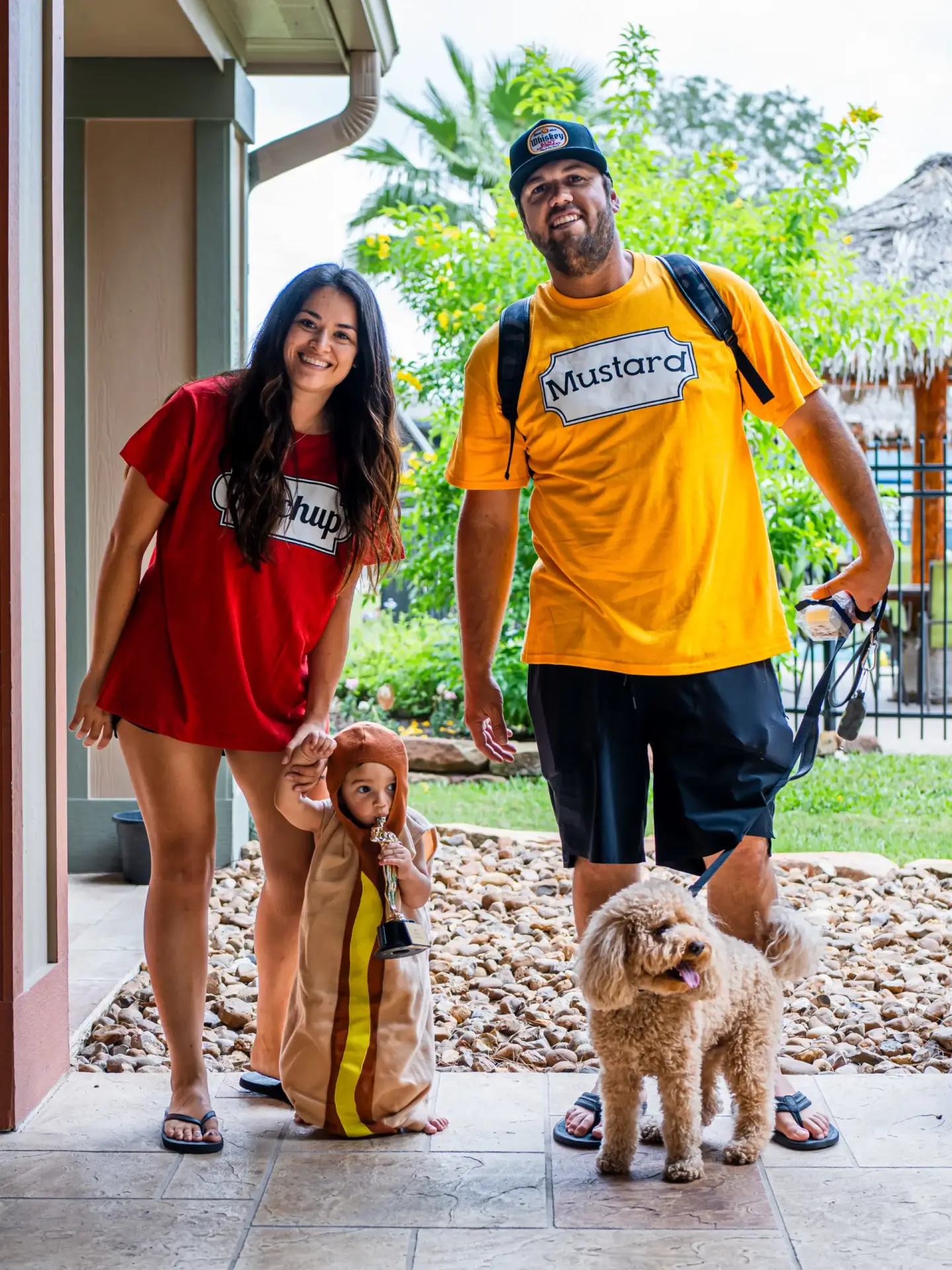 Two adults and a child in ketchup, mustard, and hot dog costumes stand at an entrance with a dog, embracing the quirky side of RV living at Rayford Crossing.