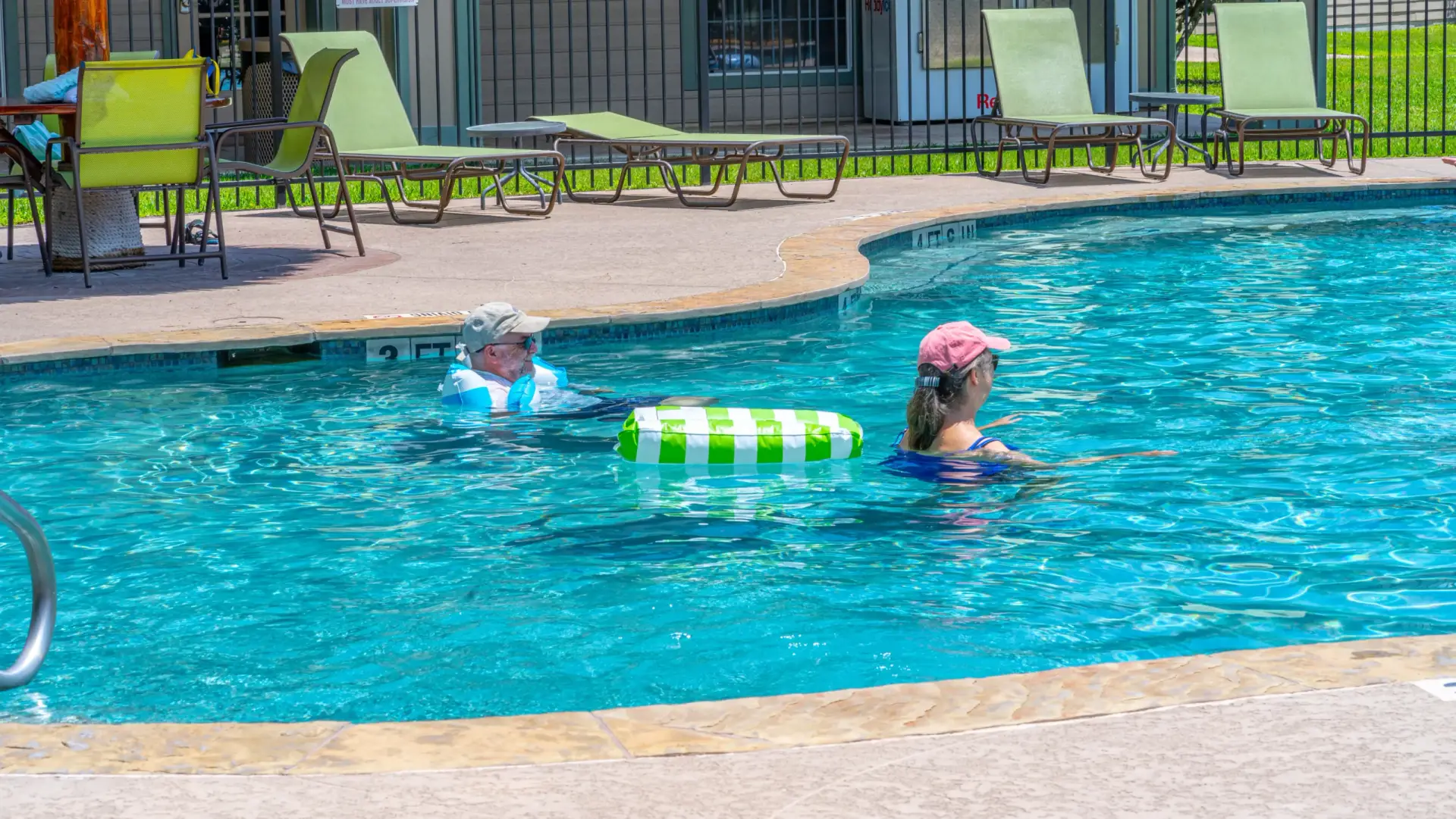 Two people wearing hats are enjoying a swim at Rayford Crossing, one with an inflatable green and white ring. Several lounge chairs hint at the relaxed, long-term RV living vibe in the background.