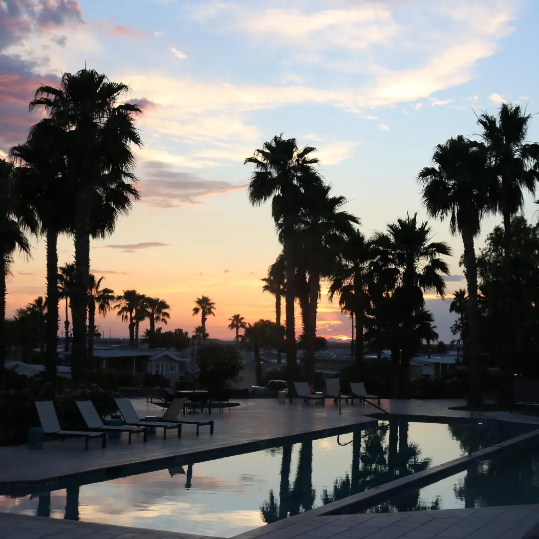 Sunset view over a swimming pool at Gold Canyon RV Resort, surrounded by palm trees and lounge chairs on the deck, creating a winter bliss scene with colorful clouds painting the sky.