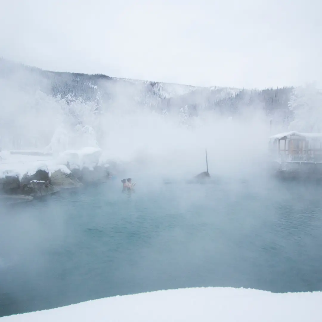 A person relaxes in a steaming hot spring in Durango, surrounded by snow-covered terrain and trees under a cloudy sky.