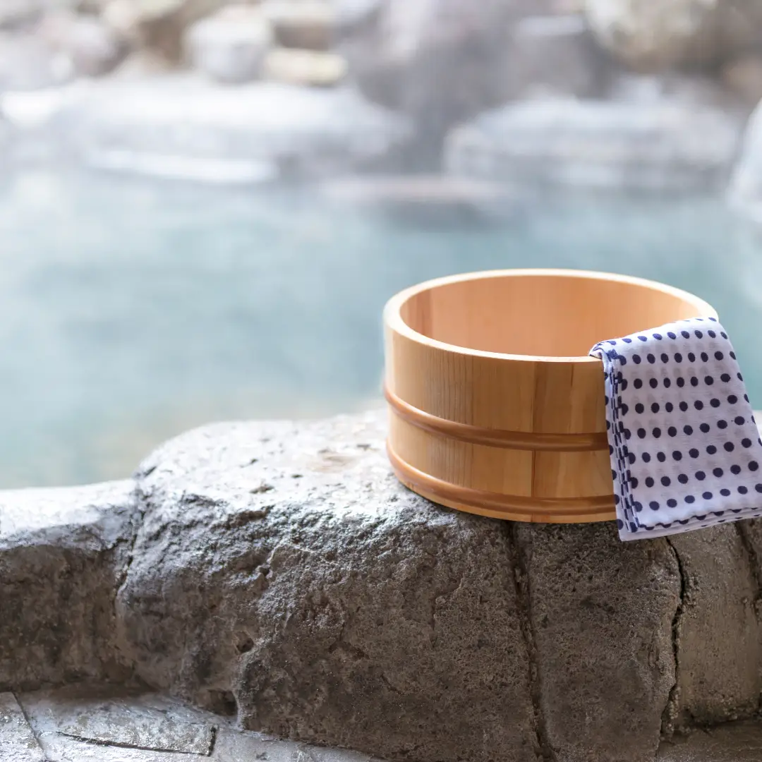 A wooden bucket with a polka-dotted towel rests on a stone near a misty hot spring, inviting you to enjoy one of the perfect spring activities. Explore Durango and immerse yourself in this serene experience.