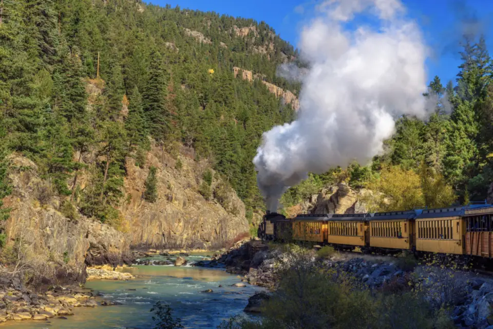 A steam train travels through the forested mountainous region of Durango, Colorado, alongside a river, emitting white smoke under a clear blue sky—one of the exhilarating summer activities to experience in the area.