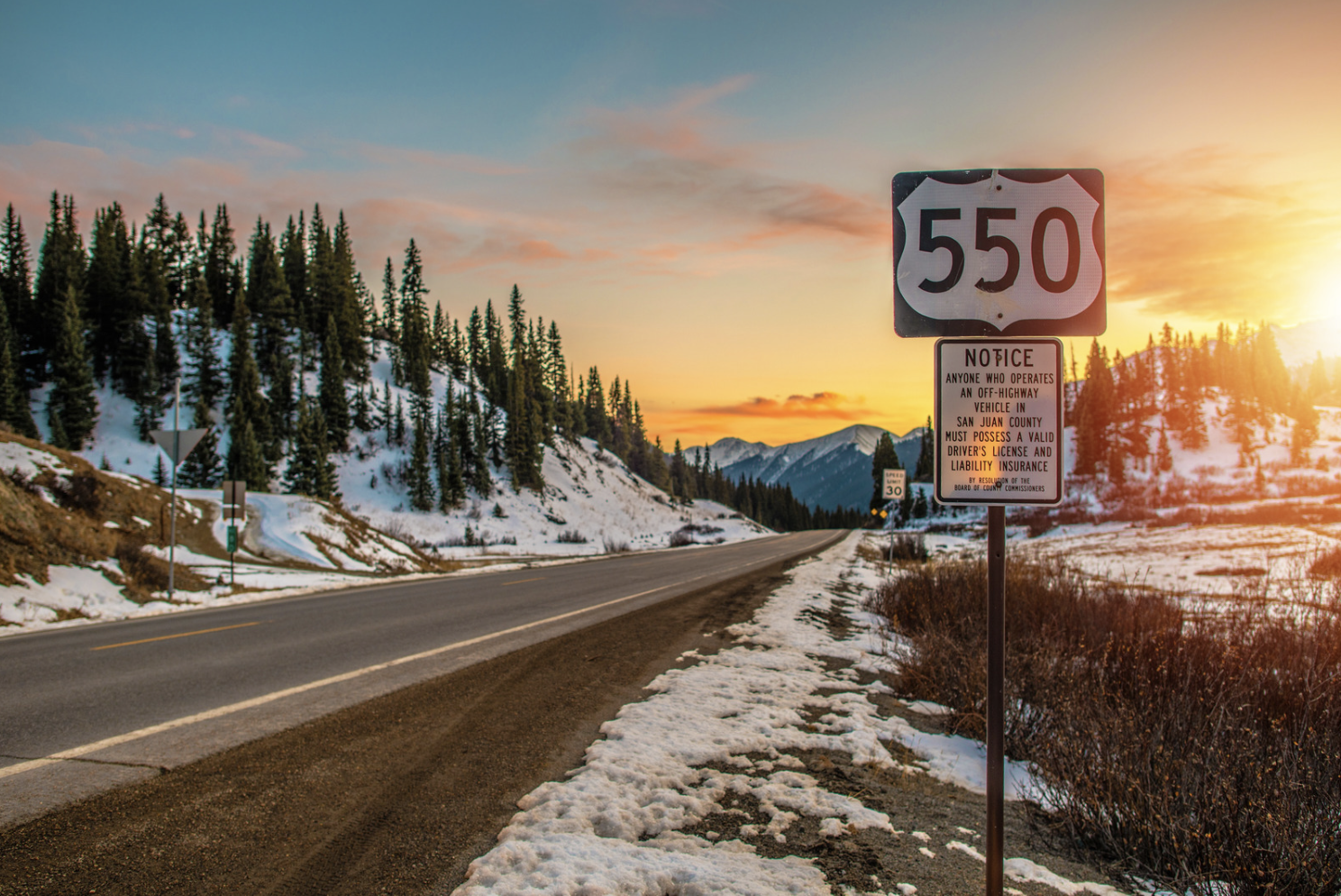 A road sign for Route 550 stands against a snow-covered, mountainous landscape at sunset near Durango. Pines line the road, offering a serene backdrop perfect for an off-season winter RV adventure.