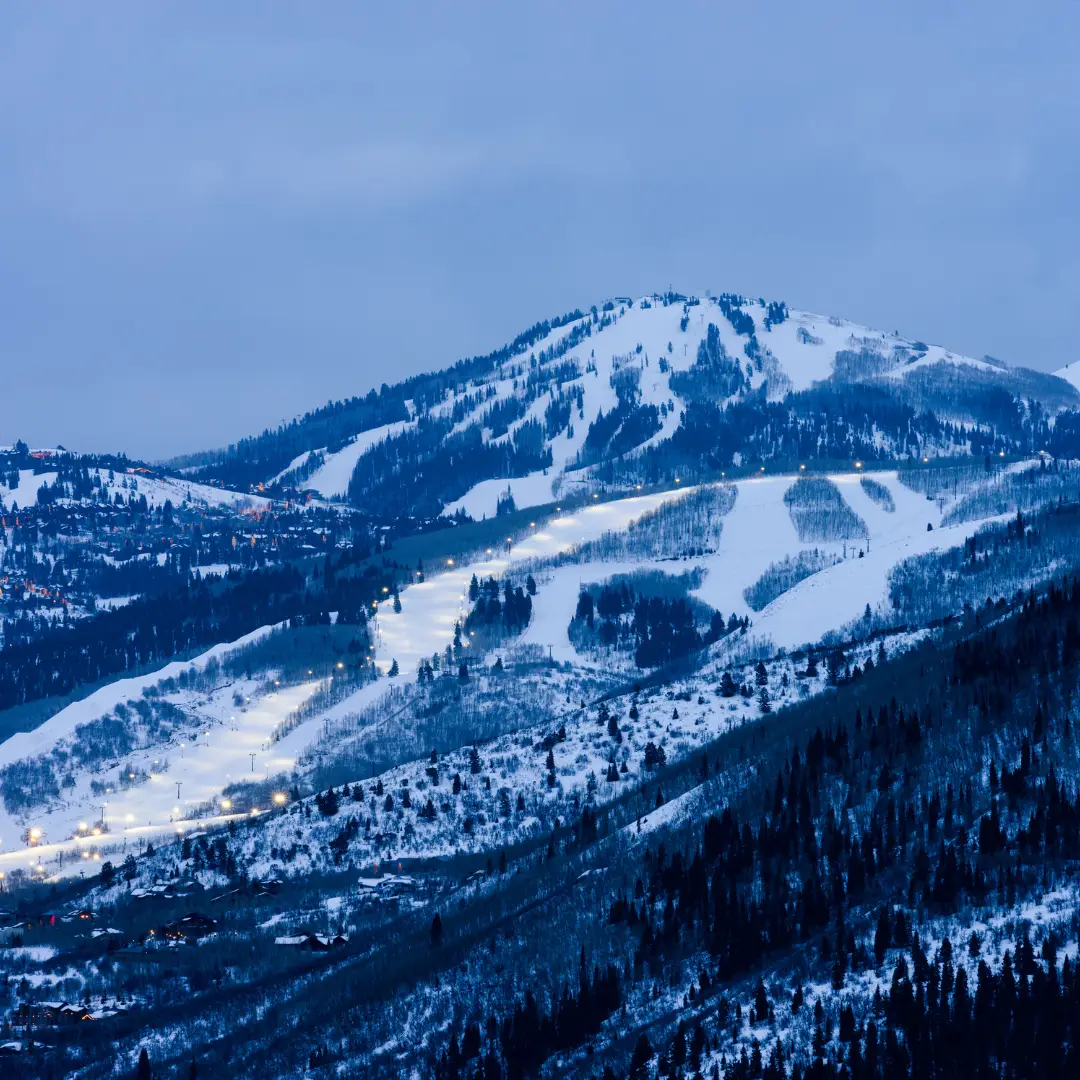 At dusk, the snow-covered mountain of Park City gleams, its ski slopes inviting enthusiasts for a winter adventure amidst trees and rugged terrain.