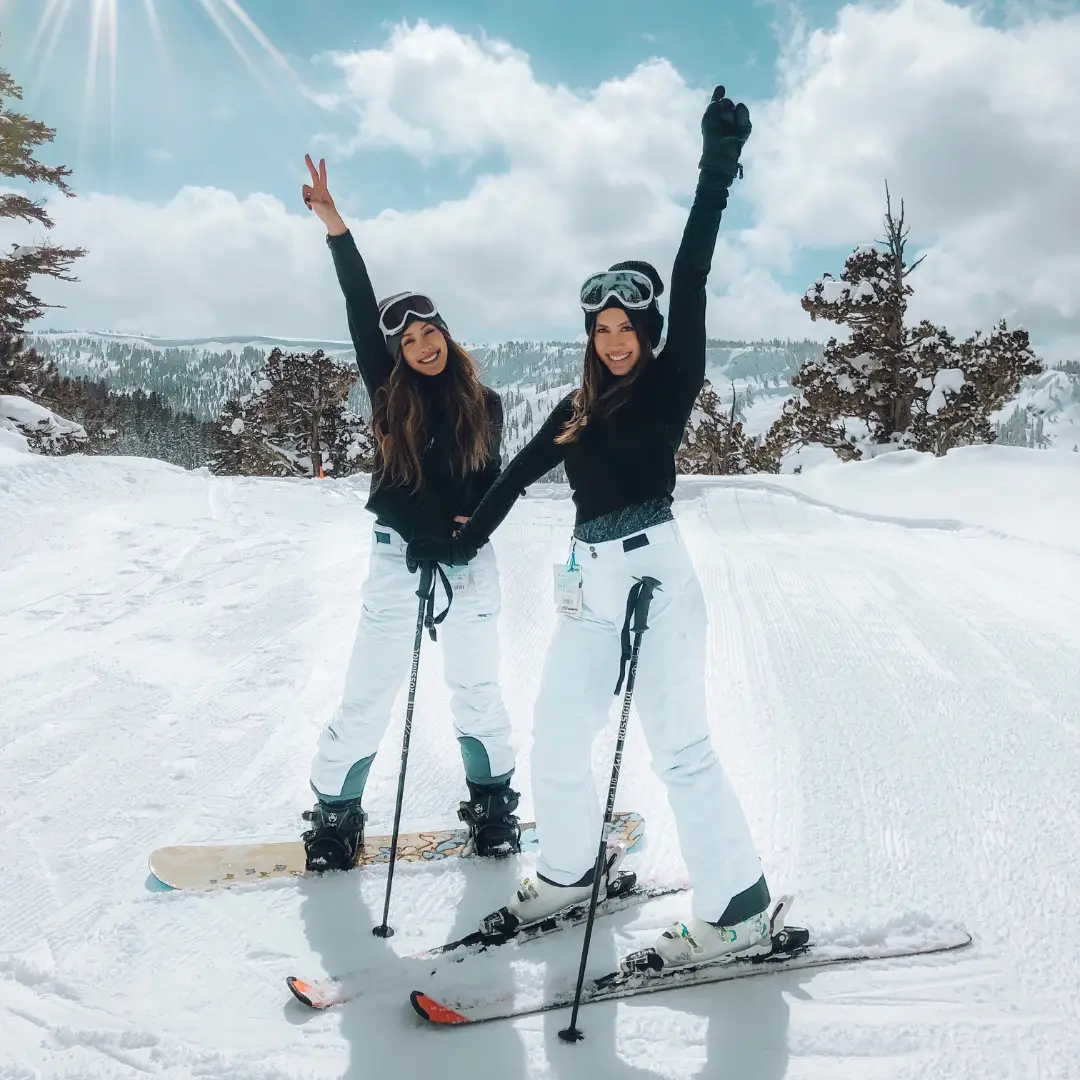 Two people in winter gear pose cheerfully with skis and snowboards on a snowy slope, embracing the thrill of a winter adventure. Park City's cloudy sky and pine trees set the backdrop for their excitement.