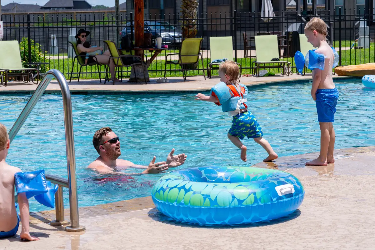 At Rayford Crossing, a child wearing a life jacket joyfully jumps into the pool towards an adult, while another in swim gear waits nearby. An inflatable ring floats lazily as people savor their RV getaway, relaxing under umbrellas and embracing the chance to fall into comfort.