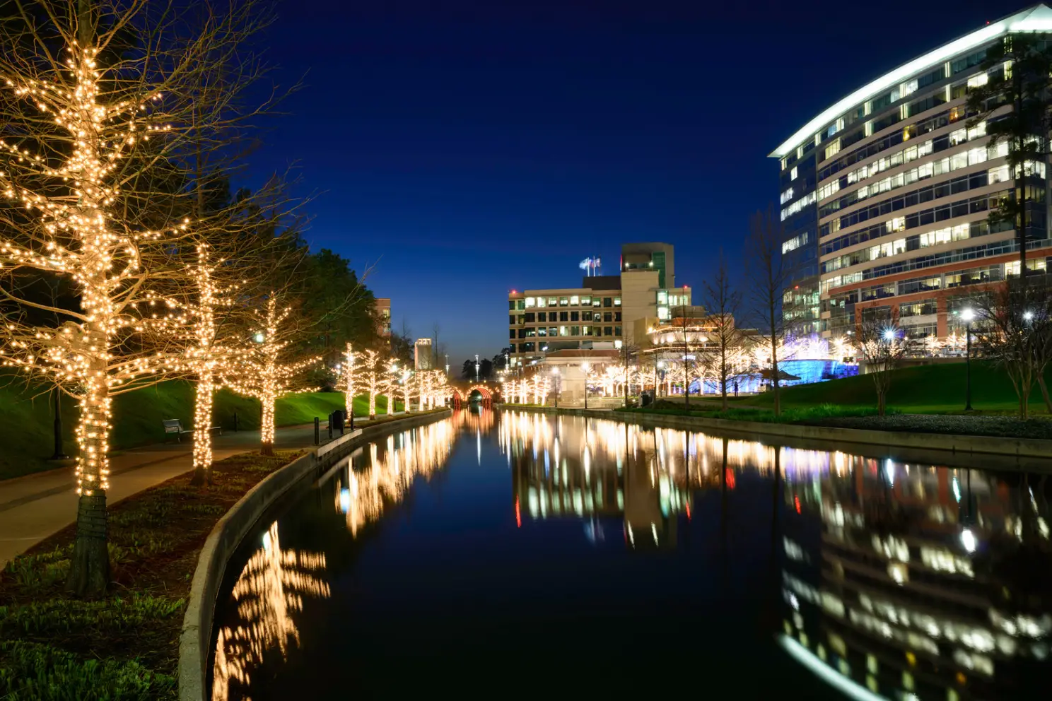 A night scene of Rayford Crossing canal reflects buildings and trees adorned with white string lights, creating a picturesque backdrop perfect for a fall into comfort RV getaway.