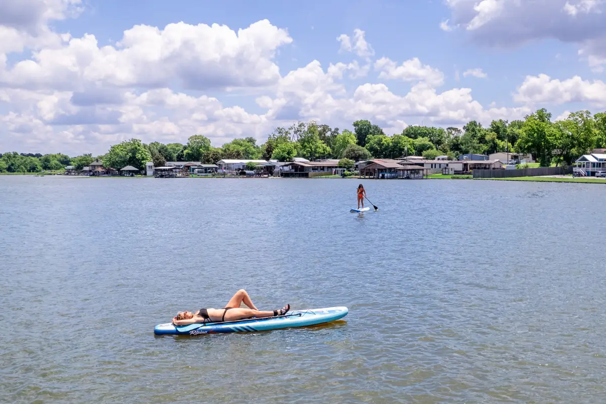 Two people enjoy the serenity of Lake Conroe: one lies on a paddleboard, while the other stands and paddles. Houses dot the water’s edge, visible under a partly cloudy sky.