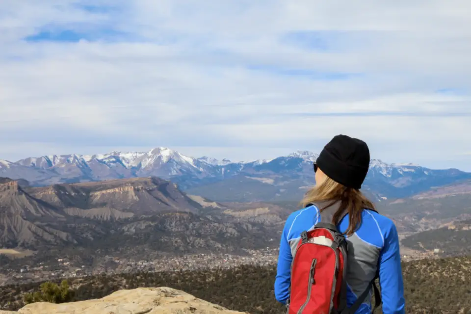 Person with a red backpack and blue jacket overlooks the rugged Durango mountainous landscape under a cloudy sky, where adventure seekers relish mountain biking trails and serene fishing spots.