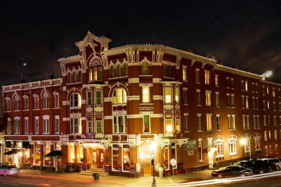 A historic, multi-story red brick hotel building in downtown Durango is illuminated at night, its lit windows and streetlights highlighting this cultural gem. Cars are parked along the street, completing the picturesque scene.