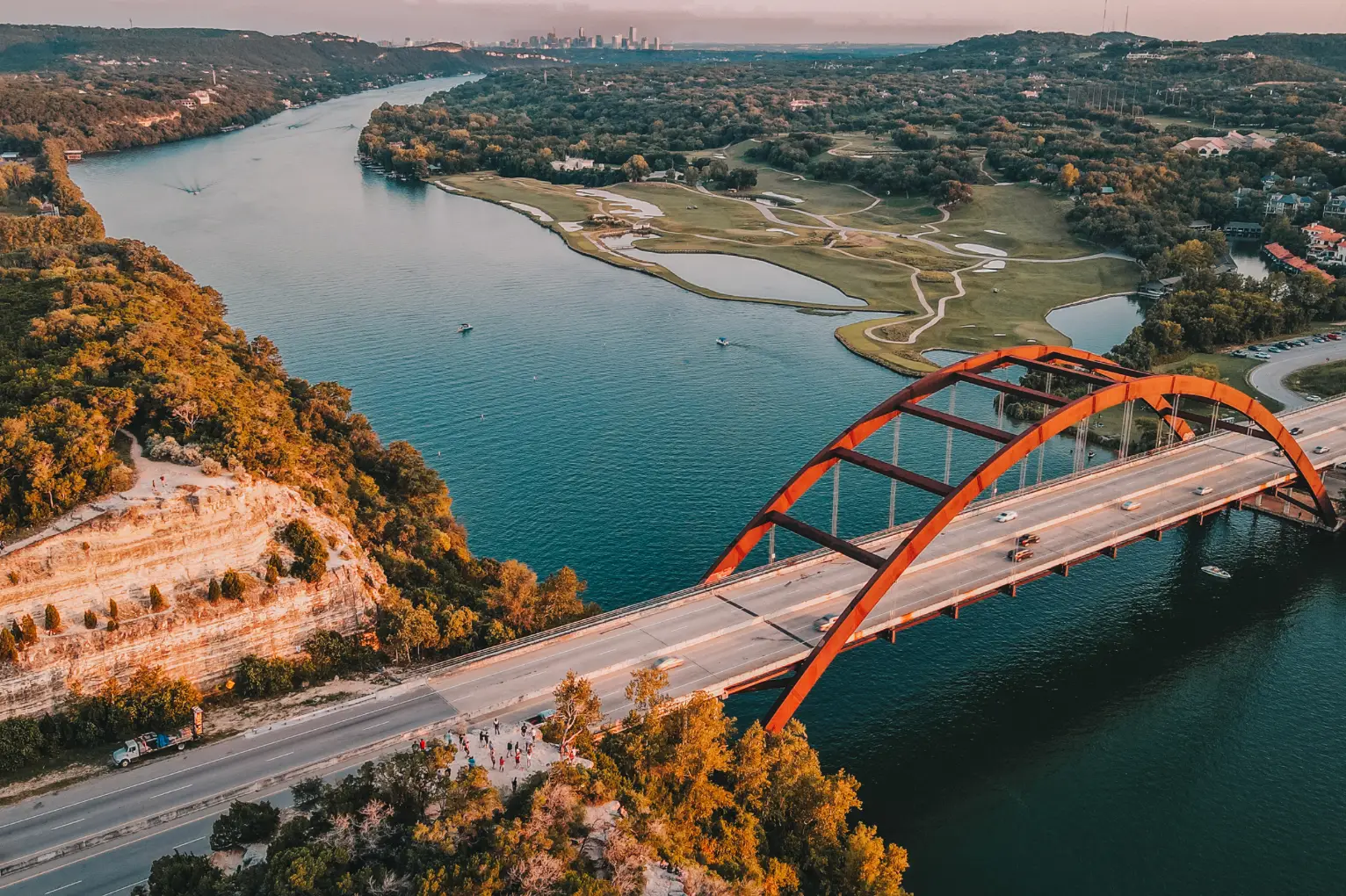Aerial view of a red arch bridge spanning a wide river, surrounded by lush greenery and distant city skyline. Cars are visible on the bridge, as locals enjoy Austin fall with vibrant foliage enhancing the scenic journey.
