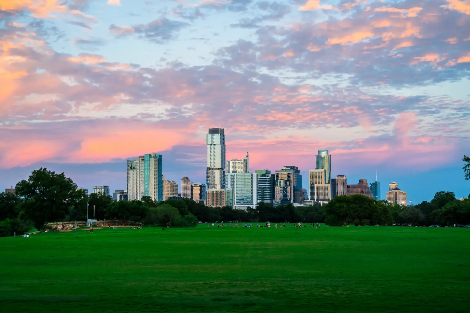 A city skyline with tall buildings is seen under a colorful sky with pink and purple clouds. In the foreground, a large green park stretches out, inviting you to enjoy fall in Austin's vibrant and picturesque setting.