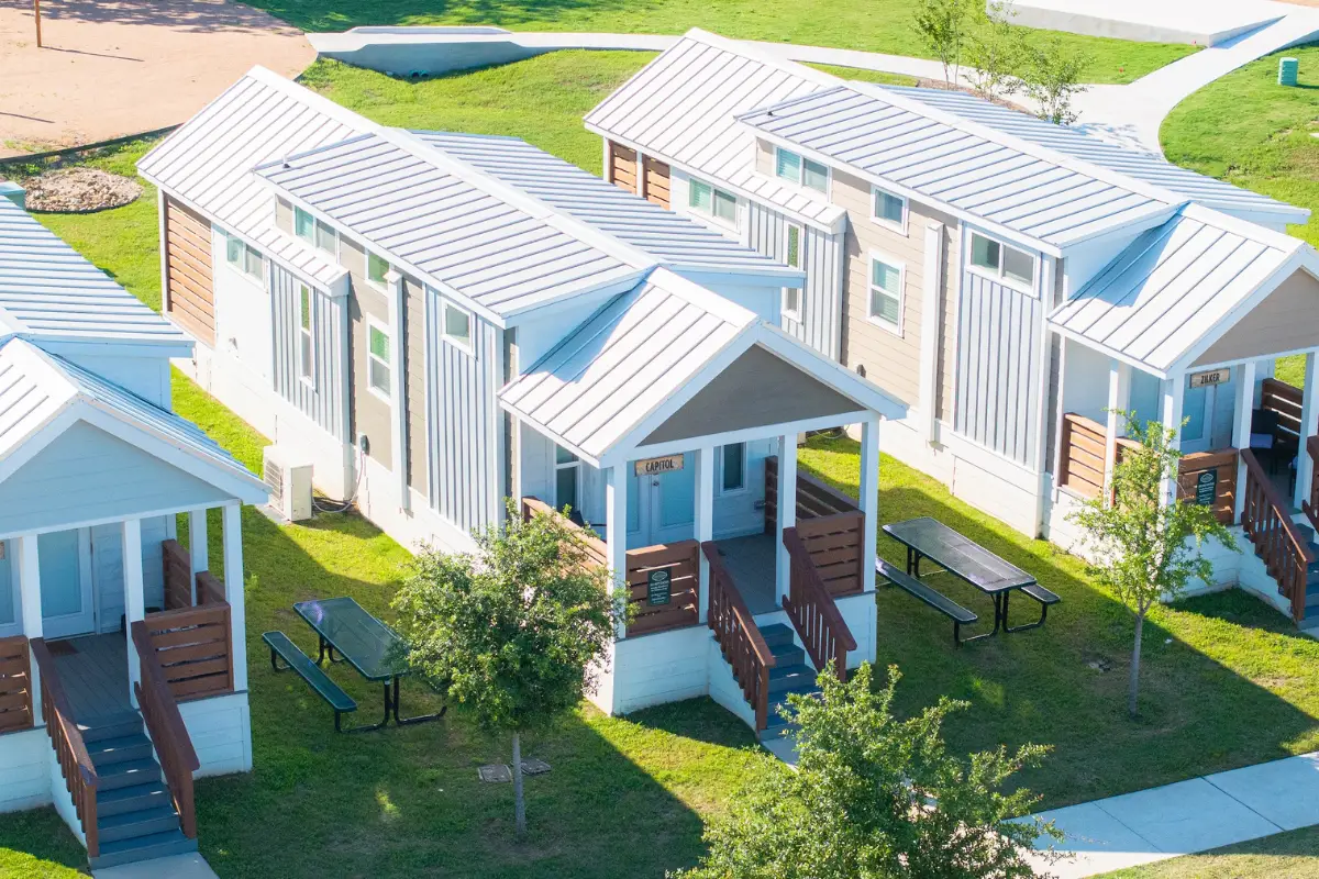 In November, an aerial view reveals three tiny houses with white roofs, nestled in the lush greenery of Austin. Each house is complemented by a picnic table set amidst the trees and grass.