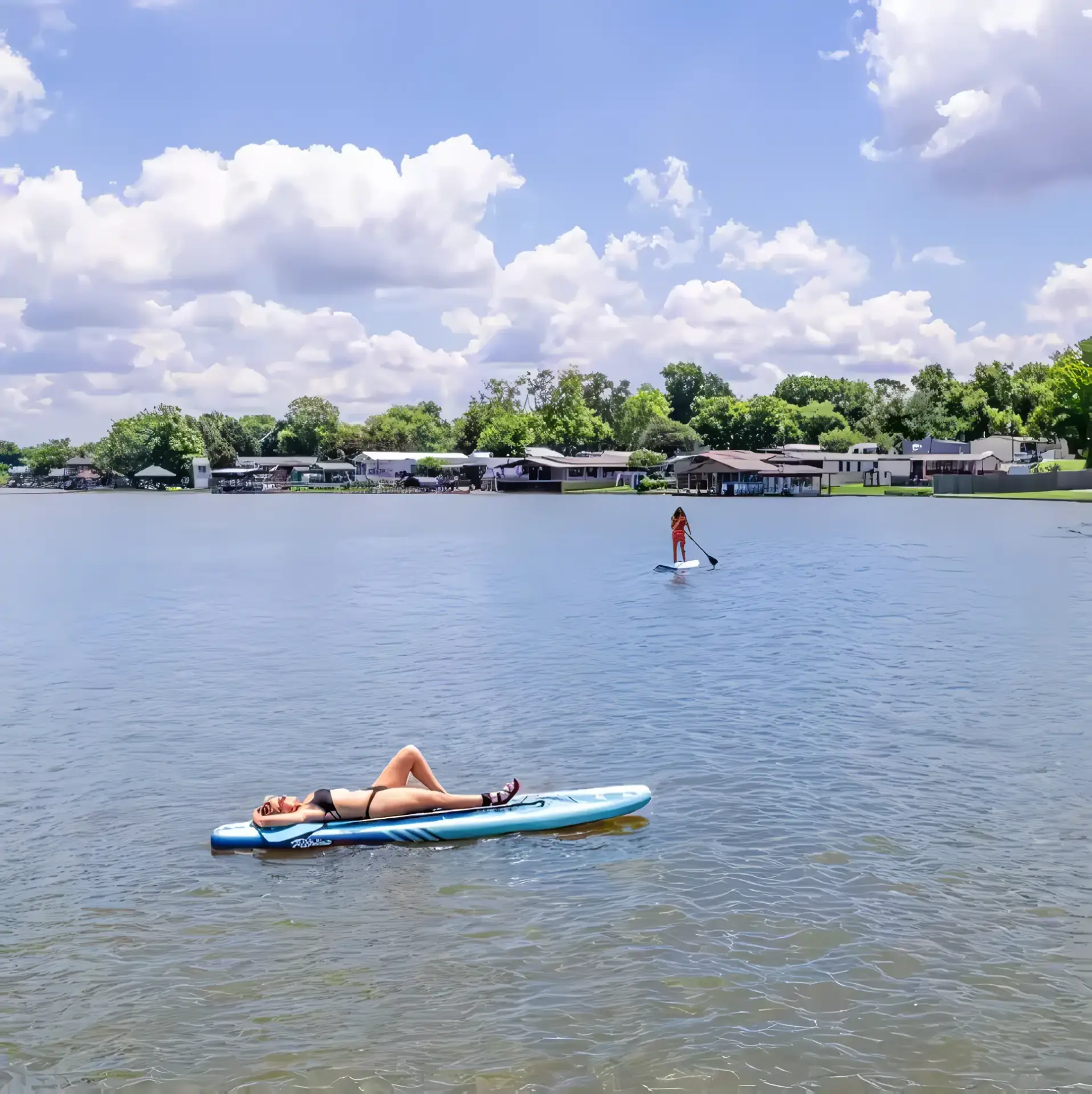 Two people enjoying the serene waters of Rayford Crossing: one lies on a paddleboard, while the other stands and paddles. Houses and trees line the shore under a partly cloudy sky, making it perfect for Family Spring Adventures.