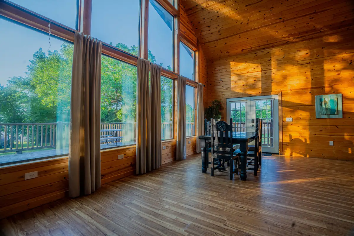 Spacious wooden room with large windows, sunlit interior, dining table, and chairs perfect for a Thanksgiving feast. Glass doors open to a balcony with views of trees and blue sky, making it an ideal lakeside retreat.