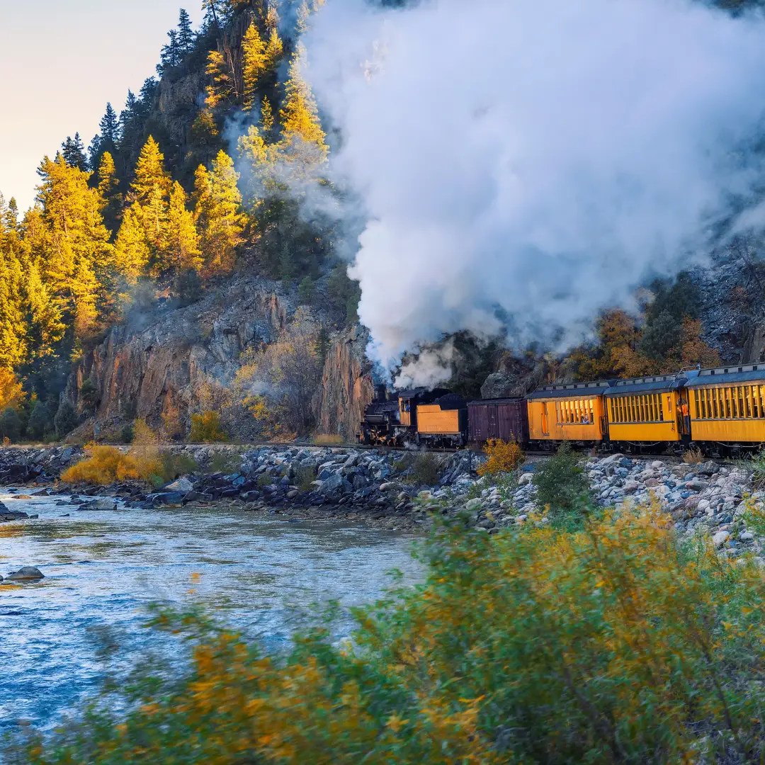 A steam train travels along a river lined with autumn trees, with smoke billowing and rocky cliffs in the background, offering a picturesque journey perfect to explore Durango's captivating landscapes.