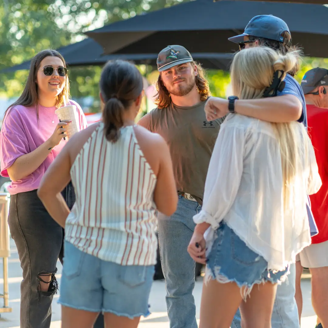 A group of people stands outdoors near Lake Conroe, chatting and enjoying the fresh air. Some wear casual clothing, perfect for their Spring Adventures in Willis, TX, with an umbrella set up in the background for added comfort.