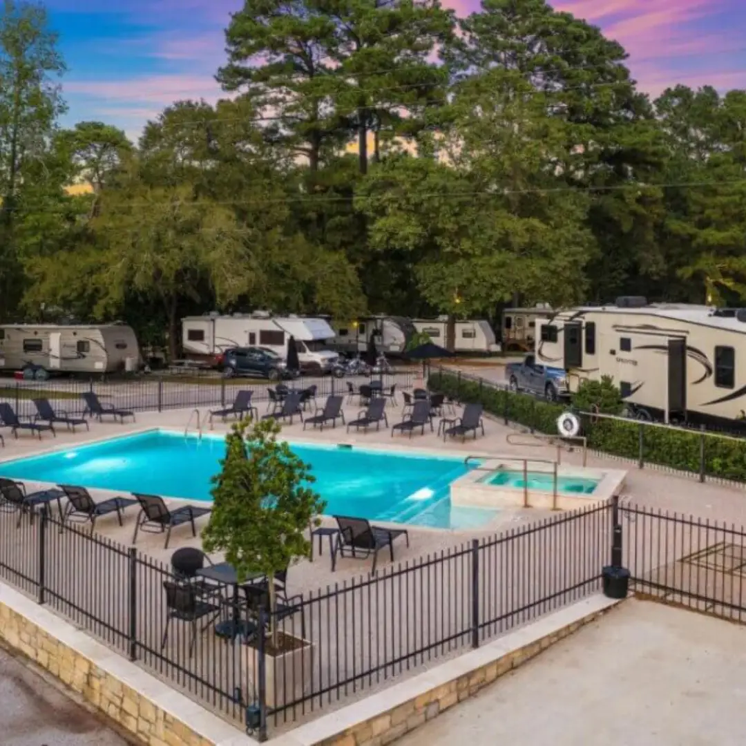 The swimming pool, surrounded by lounge chairs and tables within a fenced area, offers a perfect retreat. Several RVs are parked nearby, with trees under a colorful sky reminiscent of Spring Adventures at Lake Conroe near Willis TX.