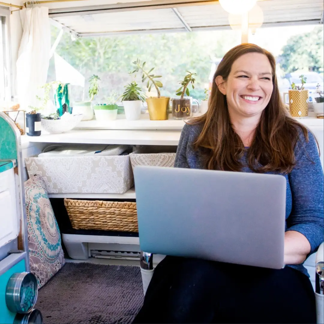 In a cozy, decorated room reminiscent of Majestic Pines, a woman smiles with a laptop on her lap, dreaming of long-term RV stays.