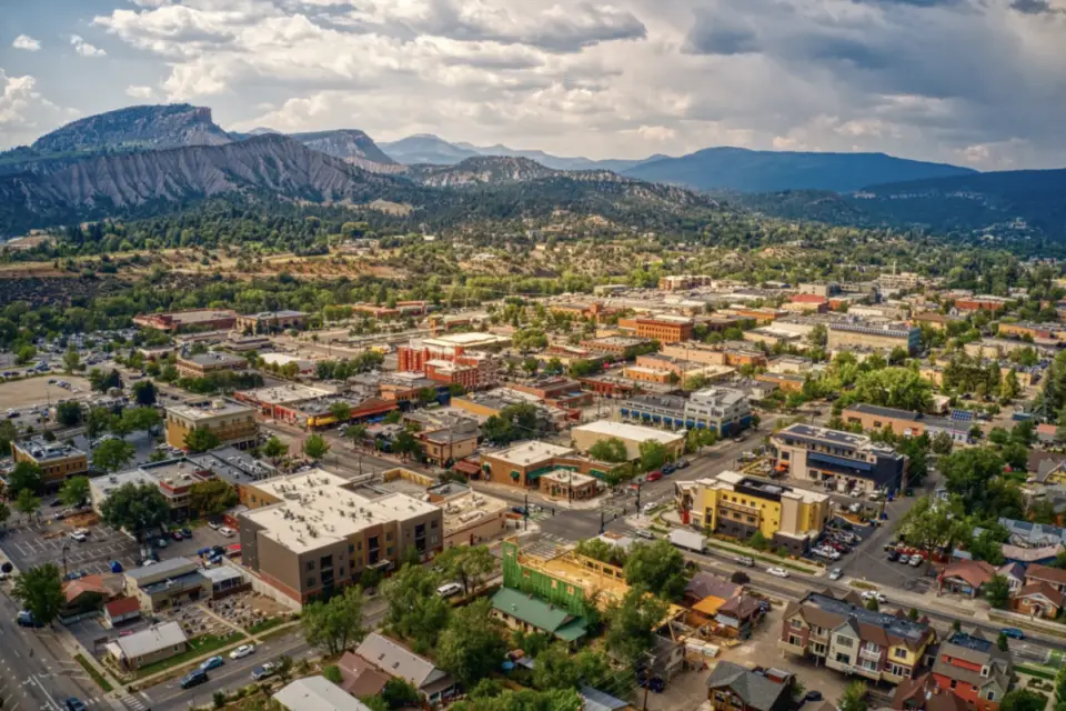 Aerial view of the town of Durango with a grid street layout nestled in a mountainous area, perfect for mountain biking, surrounded by greenery and distant hills under a partly cloudy sky.