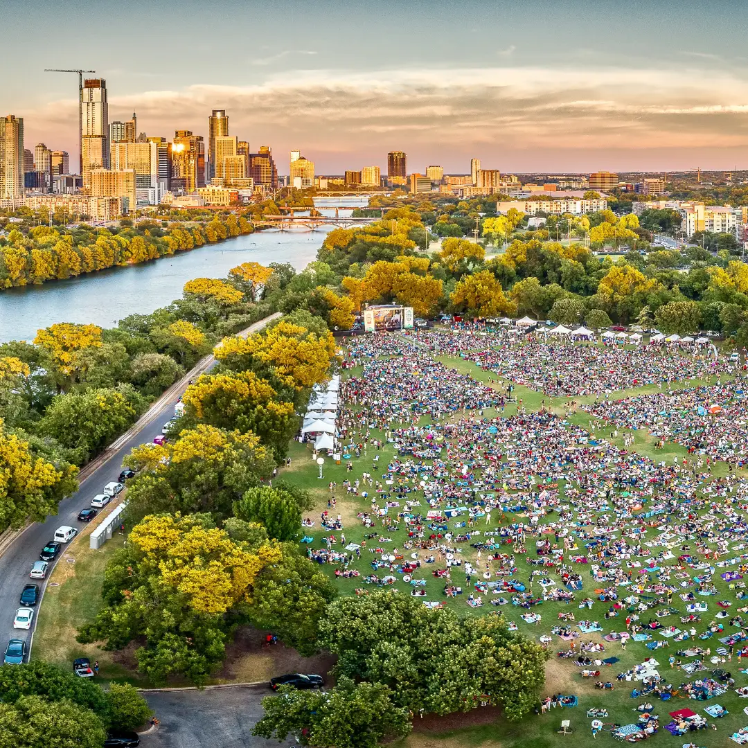 Aerial view of a large crowd gathered in a park near a river, with the Austin skyline in the background under a colorful sunset sky, hinting at vibrant upcoming events for Spring 2025.