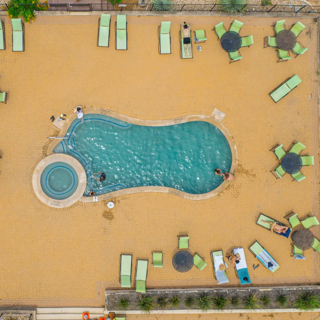 Aerial view of a bean-shaped pool with an adjacent hot tub in Austin, Texas. Several people are lounging on green chairs around the pool on a beige tiled deck.