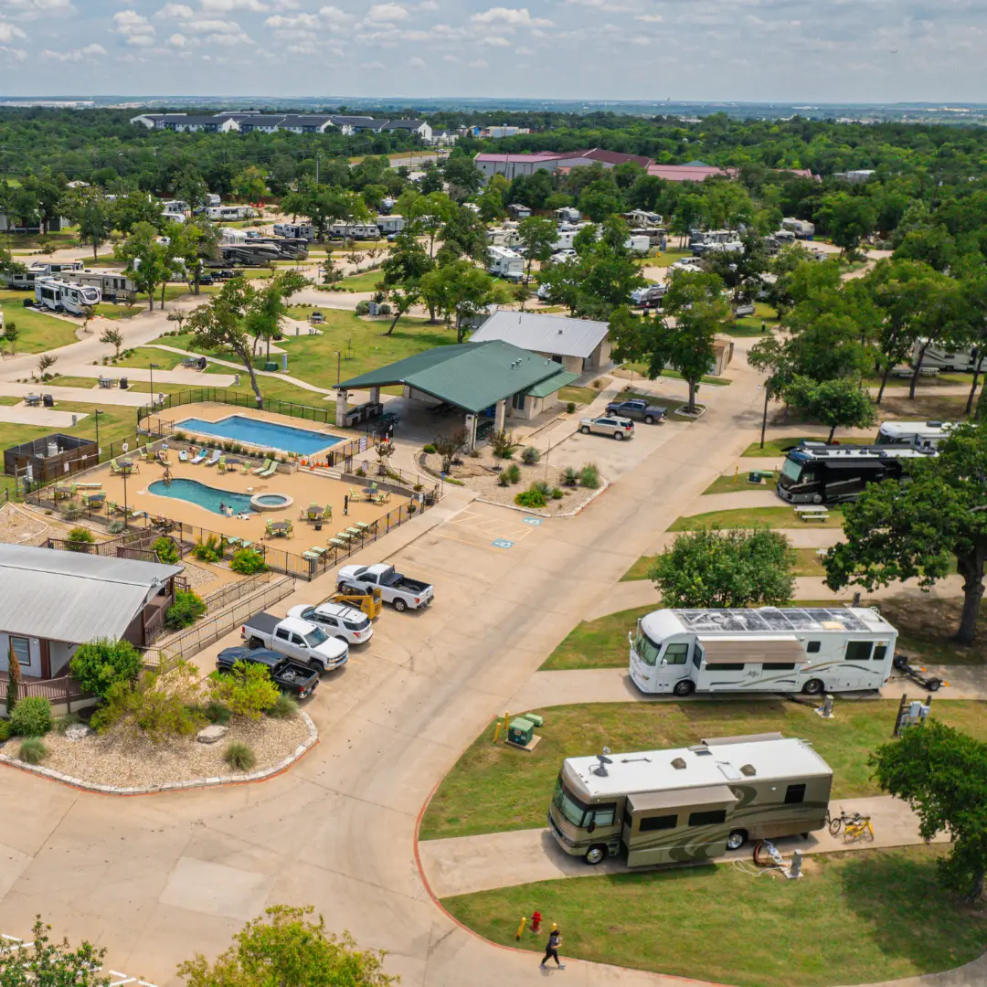 Aerial view of a perfect RV park nestled in an oak forest, featuring several RVs, cars, and a central building. There's a swimming pool, trees lining paved roads, and the landscape is expansive with greenery. Ideal for long-term RV stays.