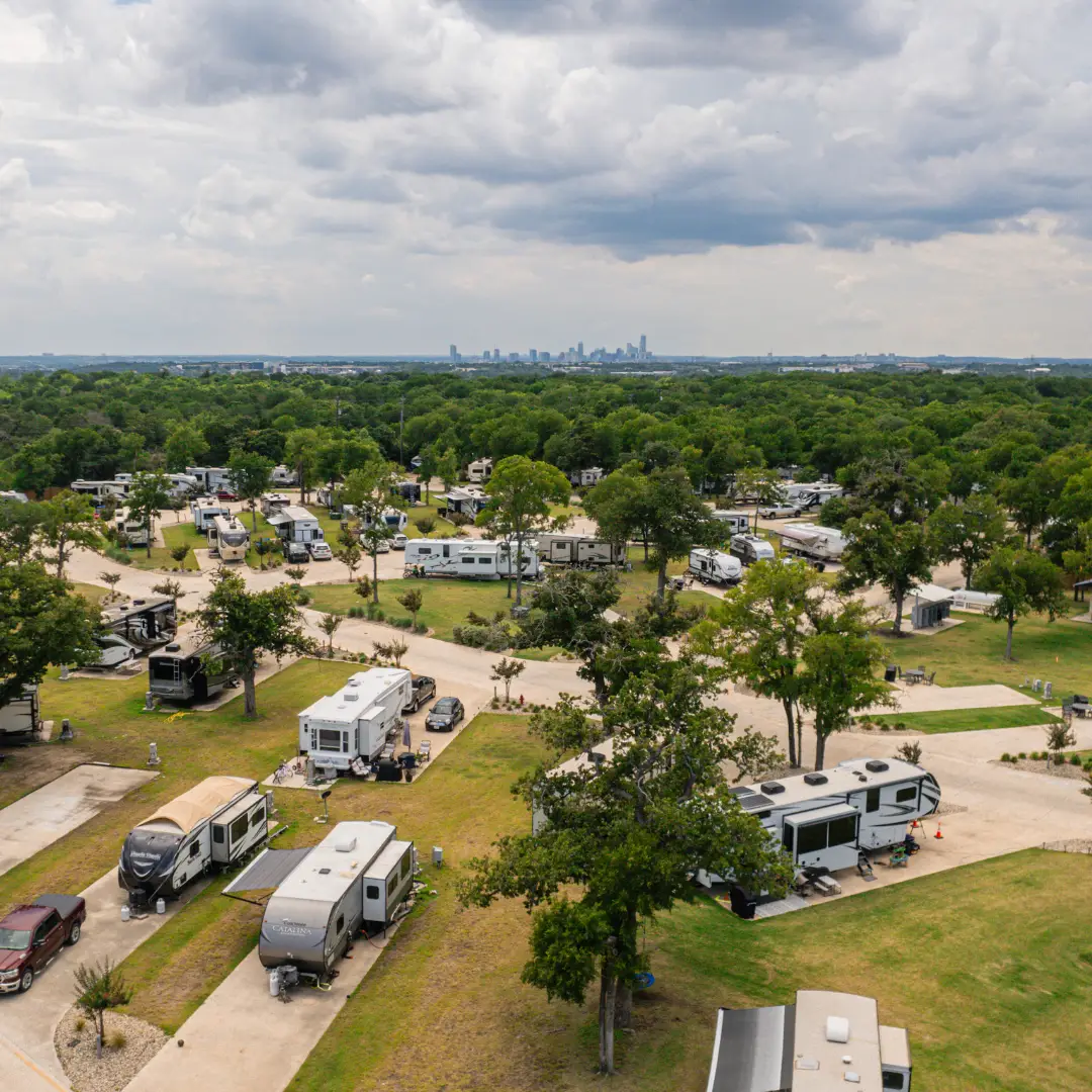 An aerial view of an Austin, Texas campground reveals numerous RVs and trailers nestled among the trees, with a city skyline visible in the background under a cloudy sky. Ideal for monthly RV living, this spot combines the benefits of nature with urban proximity.