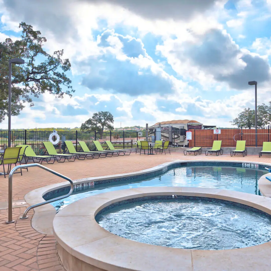 The outdoor pool area at Oak Forest is perfect for long-term RV stays, featuring lounge chairs and a hot tub under a partly cloudy sky. It's surrounded by a fence with trees in the background.