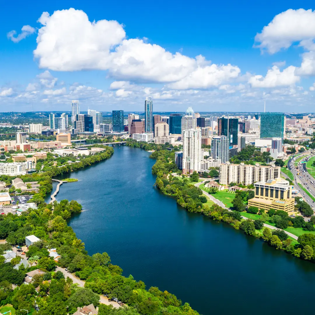 Aerial view of Austin's cityscape with a river winding through it, surrounded by trees and buildings under a partly cloudy Texas sky.