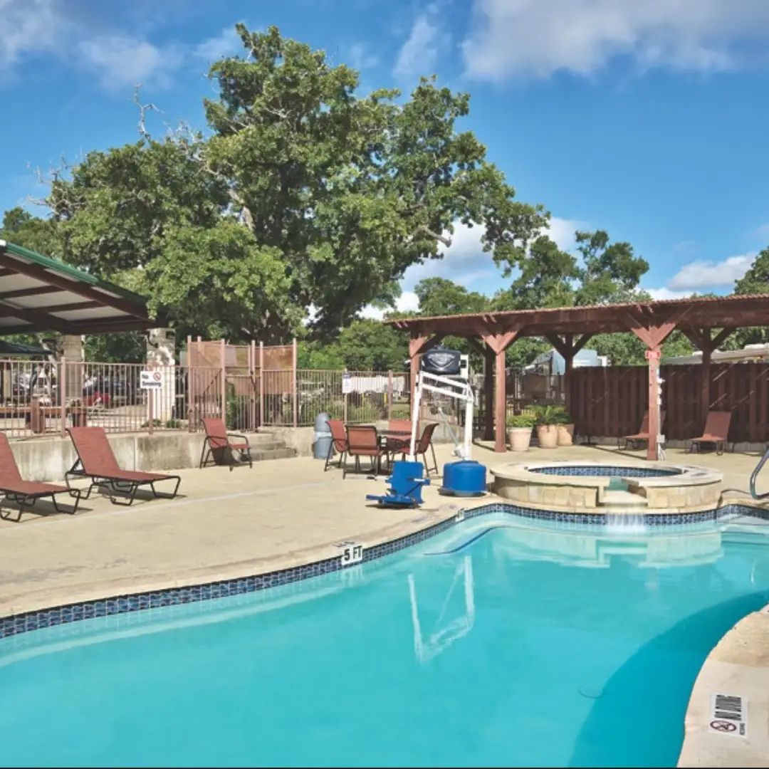 The outdoor swimming pool area boasts lounge chairs, a small hot tub, and a shaded pergola. Nestled near the serene Oak Forest, trees and a fence frame the backdrop under a partly cloudy sky, creating an ideal setting for visitors enjoying long-term RV stays.