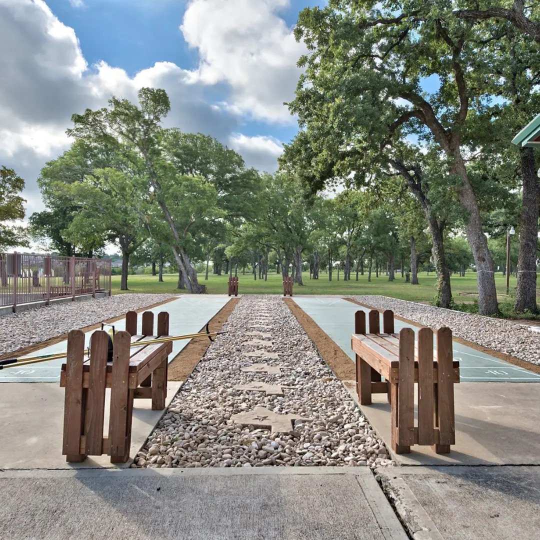 Nestled in the lush greenery of Oak Forest, an outdoor shuffleboard court awaits under a cloudy sky. Wooden benches invite relaxation as the sun peeks through, offering a serene spot for RV living enthusiasts exploring Austin, Texas.