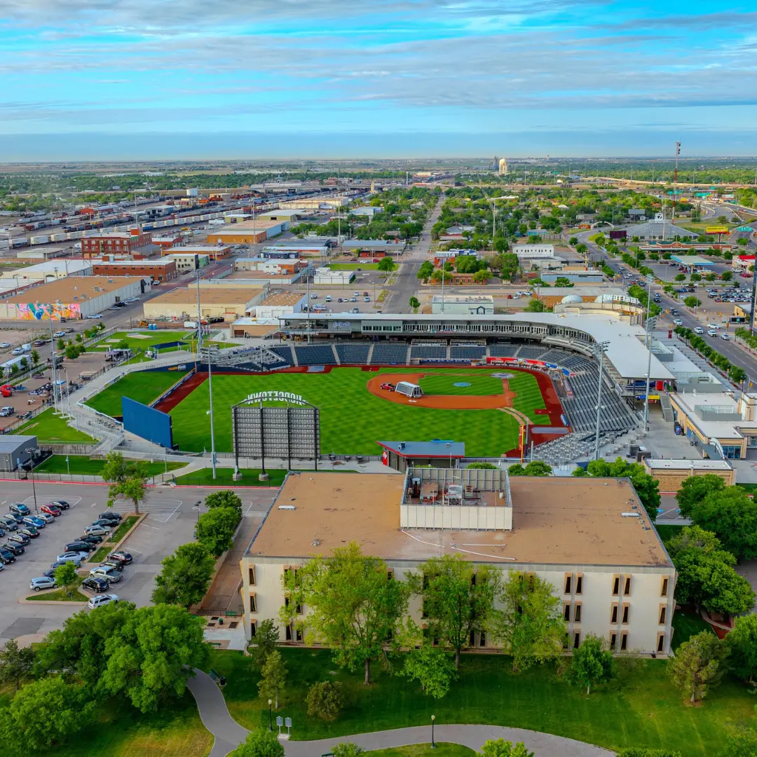 Aerial view of a baseball stadium in Amarillo, surrounded by urban buildings and lush greenery, capturing the lively atmosphere of spring events under a partly cloudy sky.