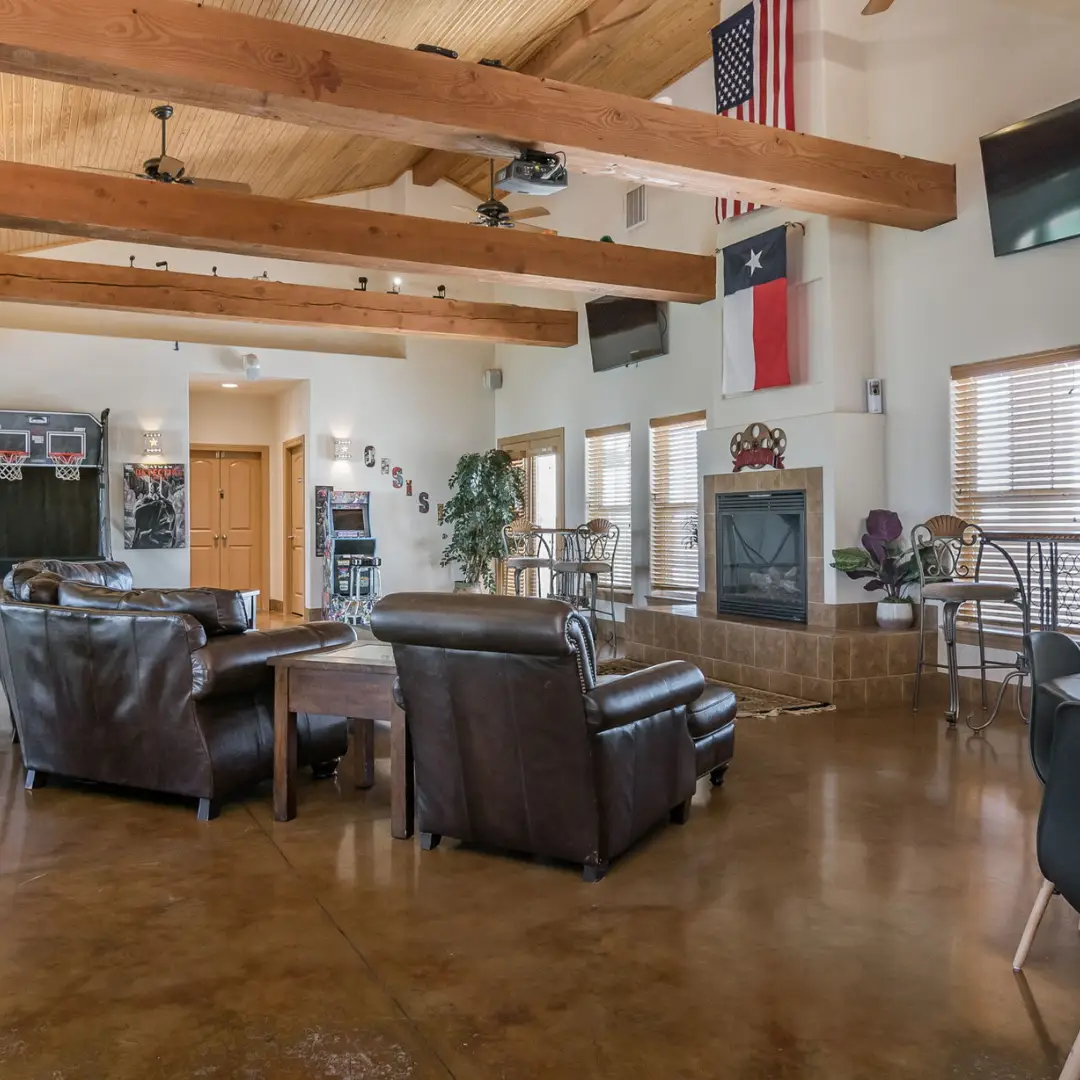 Spacious room with leather chairs, a coffee table, and a fireplace. Wooden beams run across the ceiling while American and Texas flags hang on the wall. Perfect for long-term RV travelers visiting Oasis Amarillo, there's also a basketball arcade in the corner for added fun.