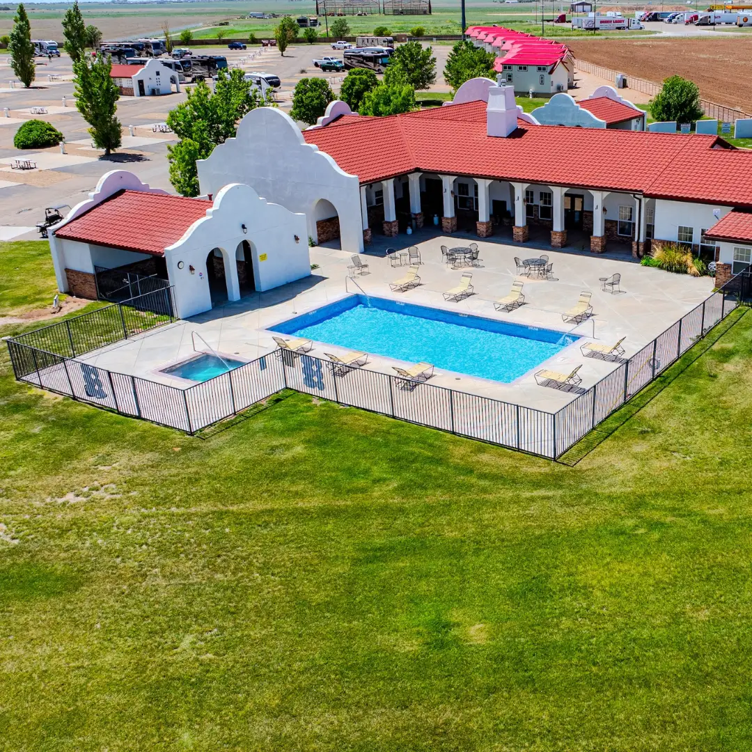 Enjoy an aerial view of a fenced outdoor swimming pool surrounded by lounge chairs, perfect for an RV overnight stop. Adjacent is a red-roofed, white building with arched architecture and a small gazebo in a grassy area, conveniently located near I-40 for easy access.