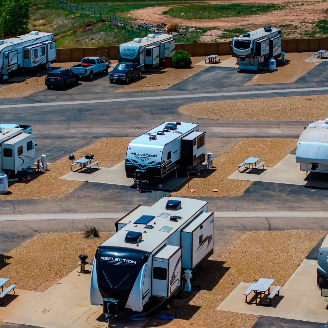 Aerial view of an RV park where trailers and RVs are parked in designated spaces, offering a home away from home. Picnic tables and parked cars enhance the cozy atmosphere. The area is surrounded by a gravel road and sparse vegetation, ideal for monthly RV stays.