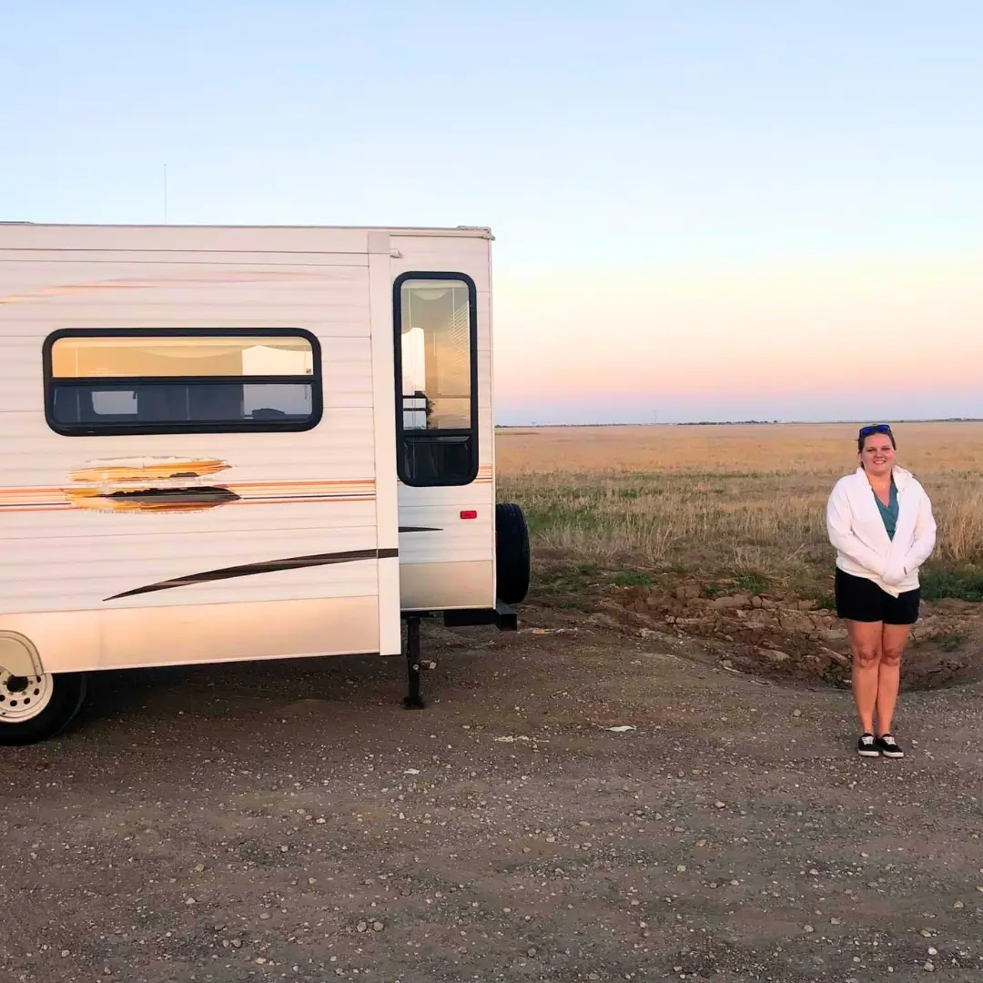 A person stands smiling next to a parked RV in an open field at sunset, enjoying the flexibility of daily RV rental.